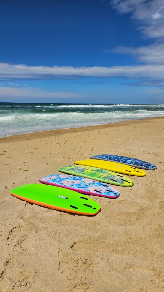 Five Ekahi soft-top surfboards in assorted colors lined up on the beach with waves in the background