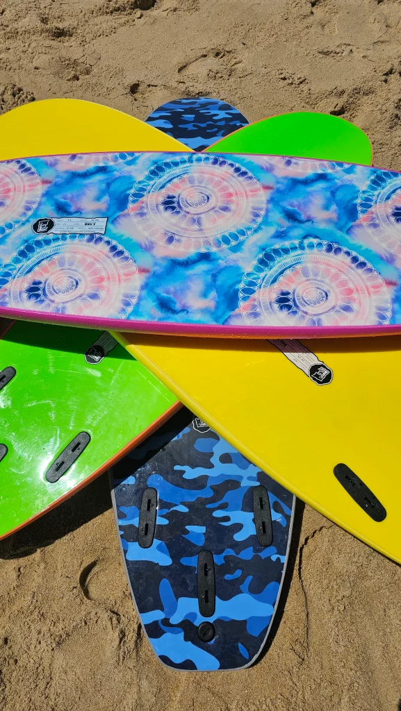 Stack of Ekahi soft-top surfboards in green, yellow, mandala tie-dye and blue camo patterns on the sand