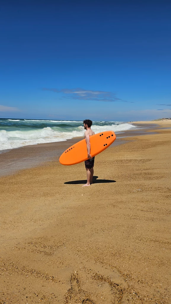 Surfer standing on the beach holding an orange Ekahi soft-top surfboard