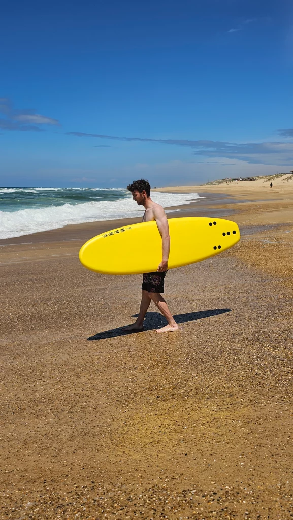 Surfer walking along the beach carrying a yellow Ekahi soft-top surfboard
