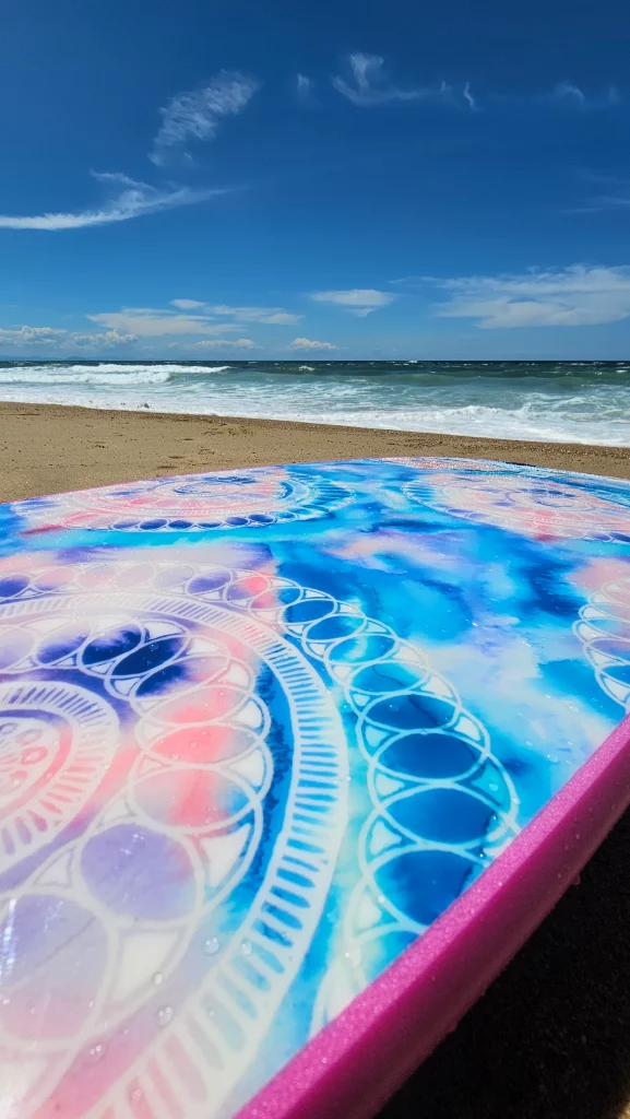 Ekahi soft-top surfboard with mandala tie-dye deck pattern and pink rail, beach in the background