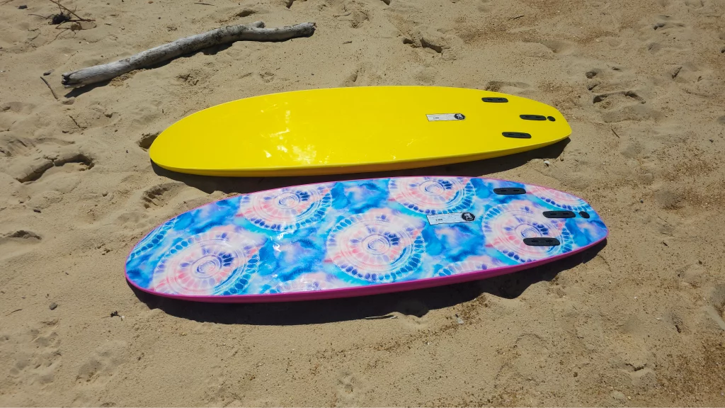 Yellow and mandala tie-dye Ekahi soft-top surfboards lying side by side on the sand
