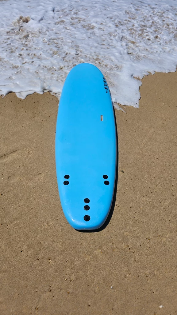 Blue Ekahi soft-top surfboard lying on the sand at the water's edge, deck view