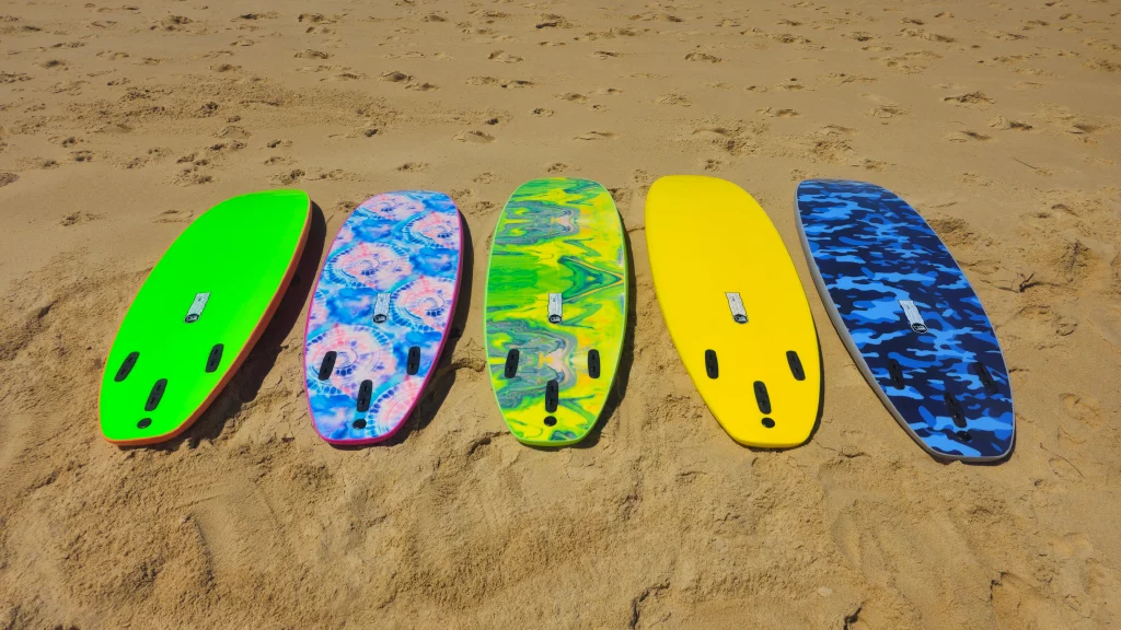Five Ekahi soft-top surfboards in green, tie-dye, marble, yellow and blue camo lying on the sand