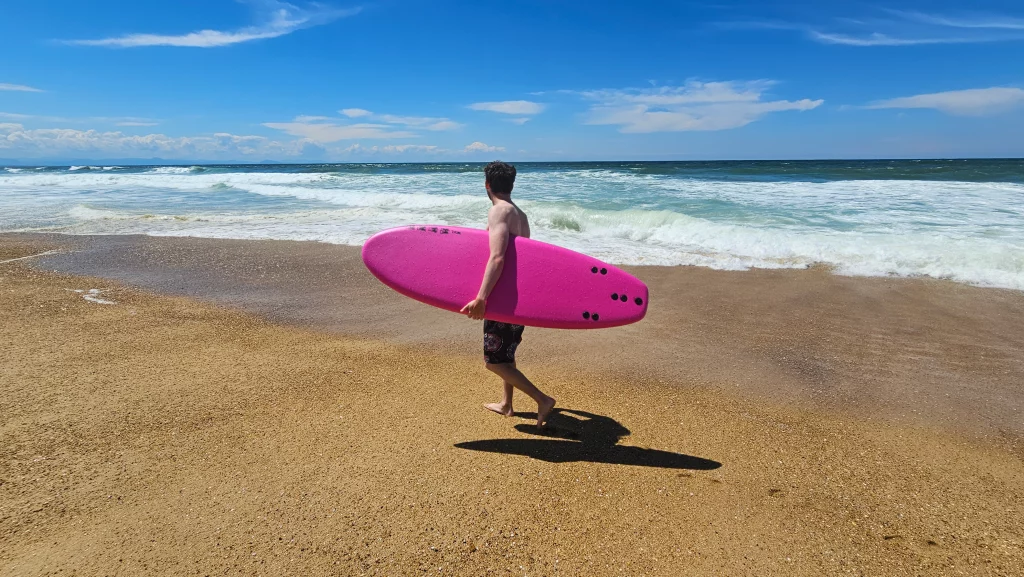 Surfer walking towards the waves carrying a pink Ekahi soft-top surfboard