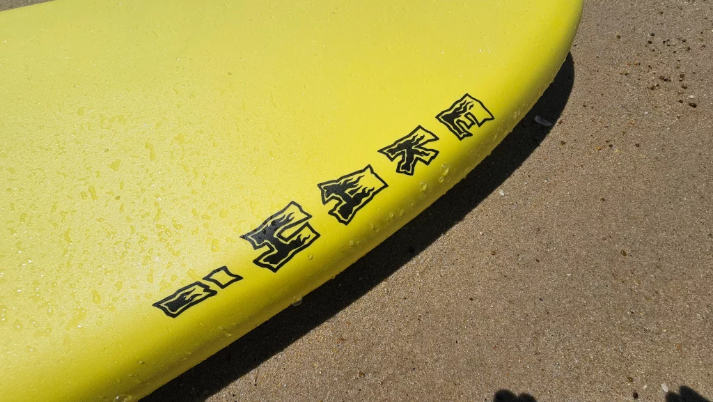 Close-up of the Ekahi logo embossed on the rail of a yellow soft-top surfboard