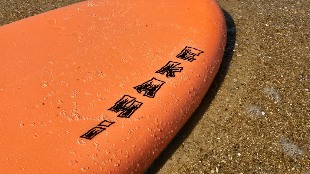 Close-up of the Ekahi logo on the deck of an orange soft-top surfboard on pebbled sand