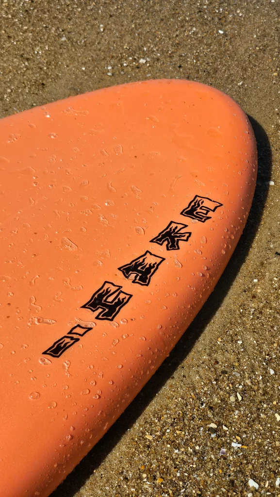Close-up of the Ekahi logo on the deck of an orange soft-top surfboard on the sand
