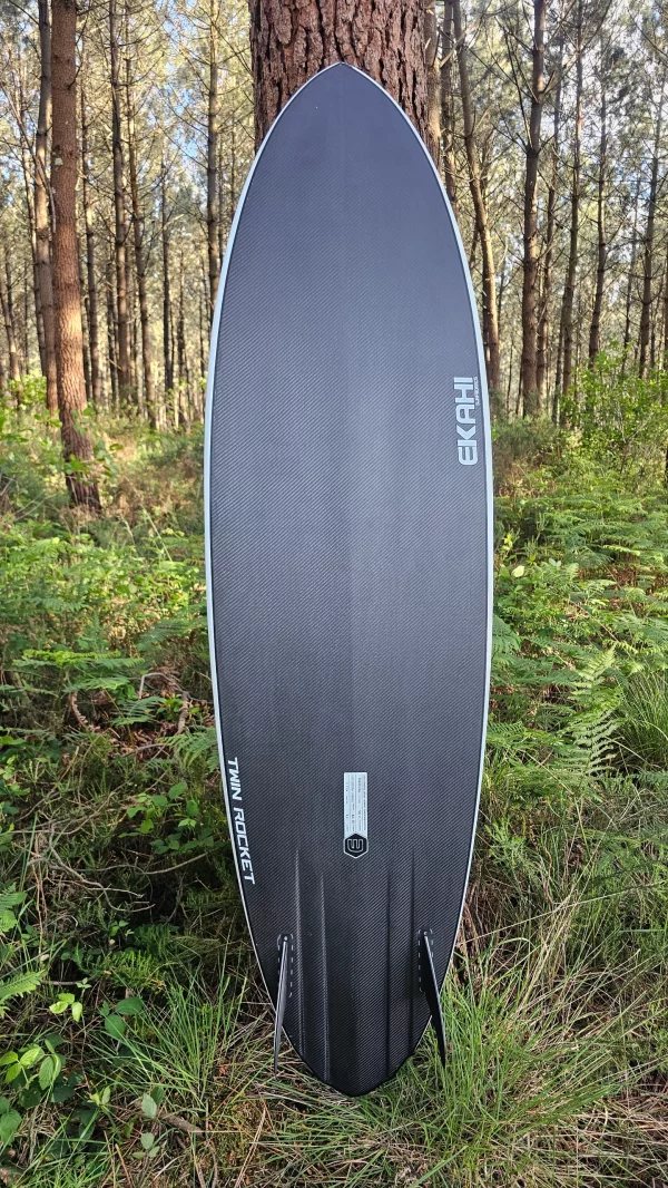 Bottom view of Ekahi white Twin Rocket carbon fiber surfboard with channels and twin fins in the forest.