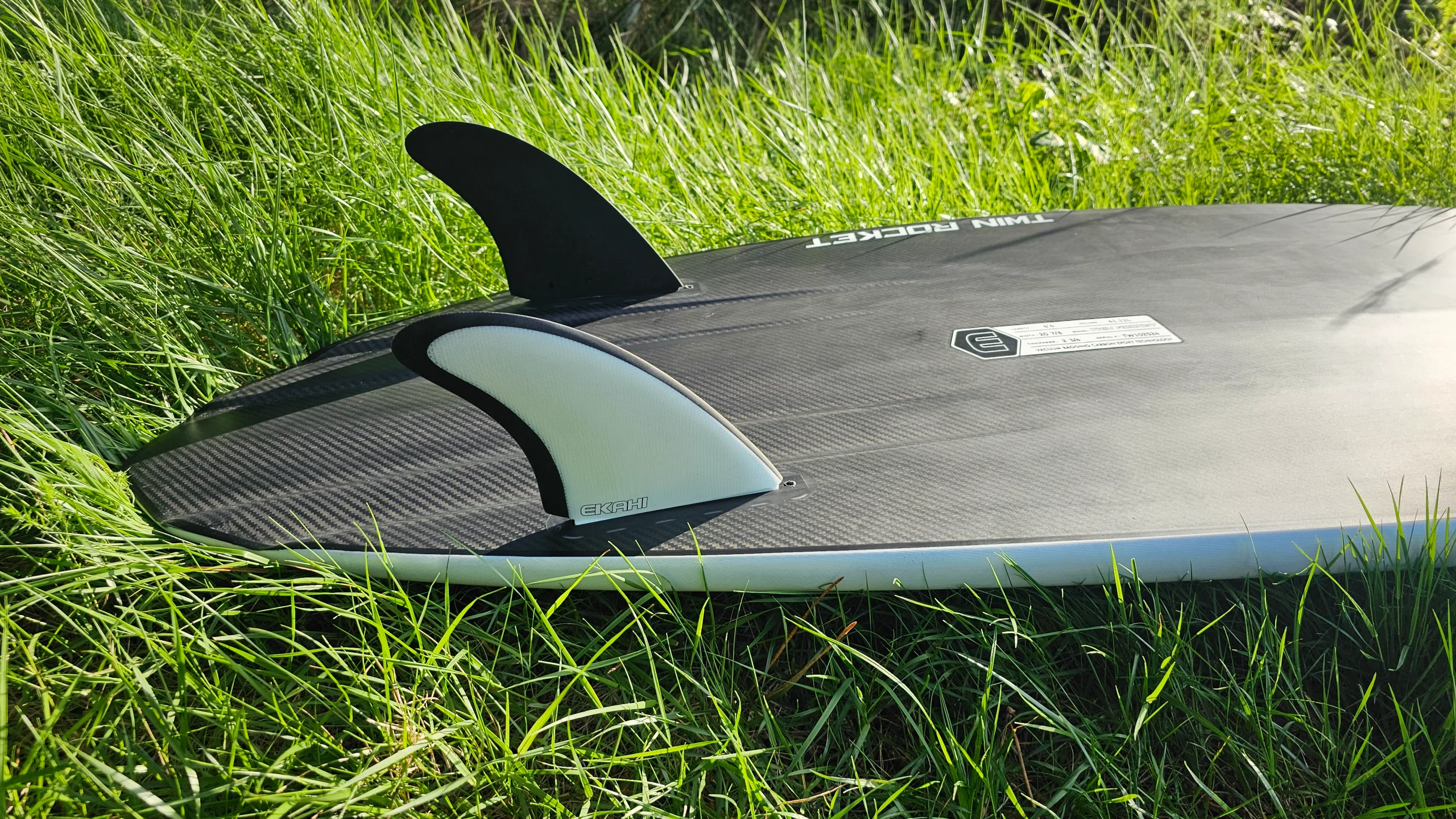 Bottom view of Ekahi white Twin Rocket carbon fiber surfboard with twin fins on the grass.
