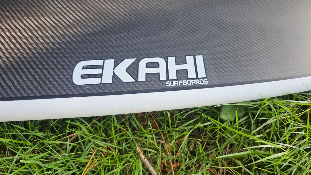 Bottom Ekahi logo view of white Twin rocket carbon fiber surfboard on the grass.