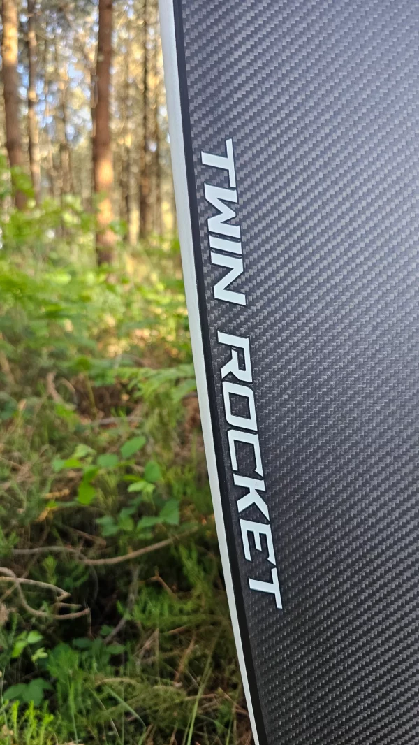 Bottom Twin rocket logo view of Ekahi white carbon fiber surfboard on the forest.