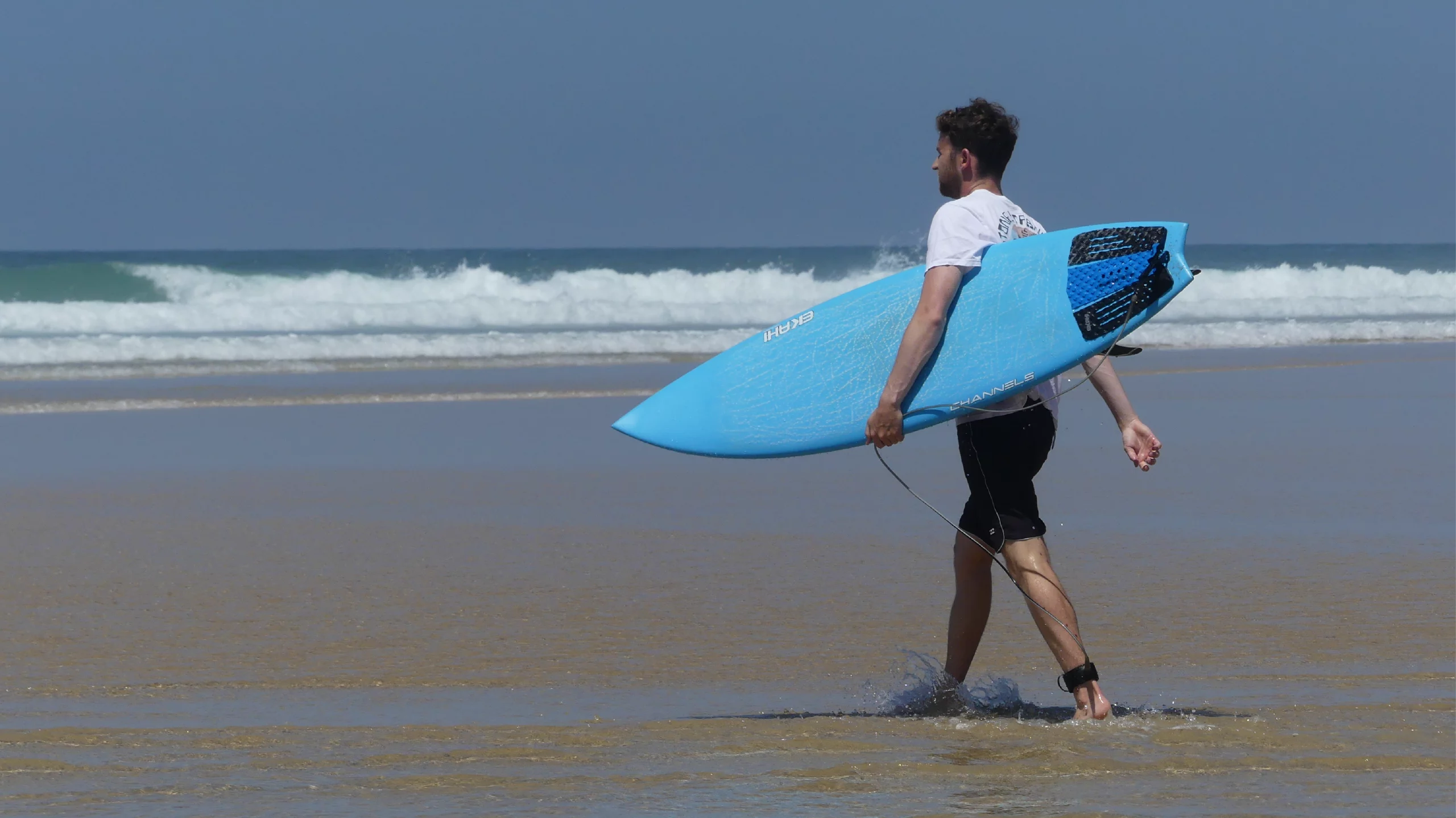Surfer walking on the beach with a blue color swallow Channels easy-rider surfboard with a squash pad.