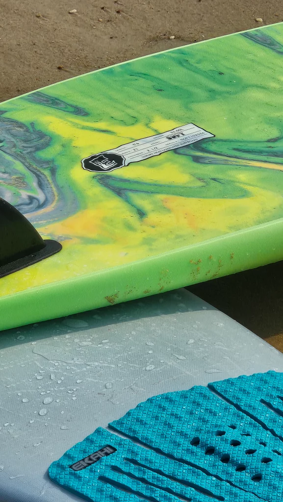 Close-up of the tail and fin on a green tie-dye Ekahi soft-top surfboard, with traction pad in foreground