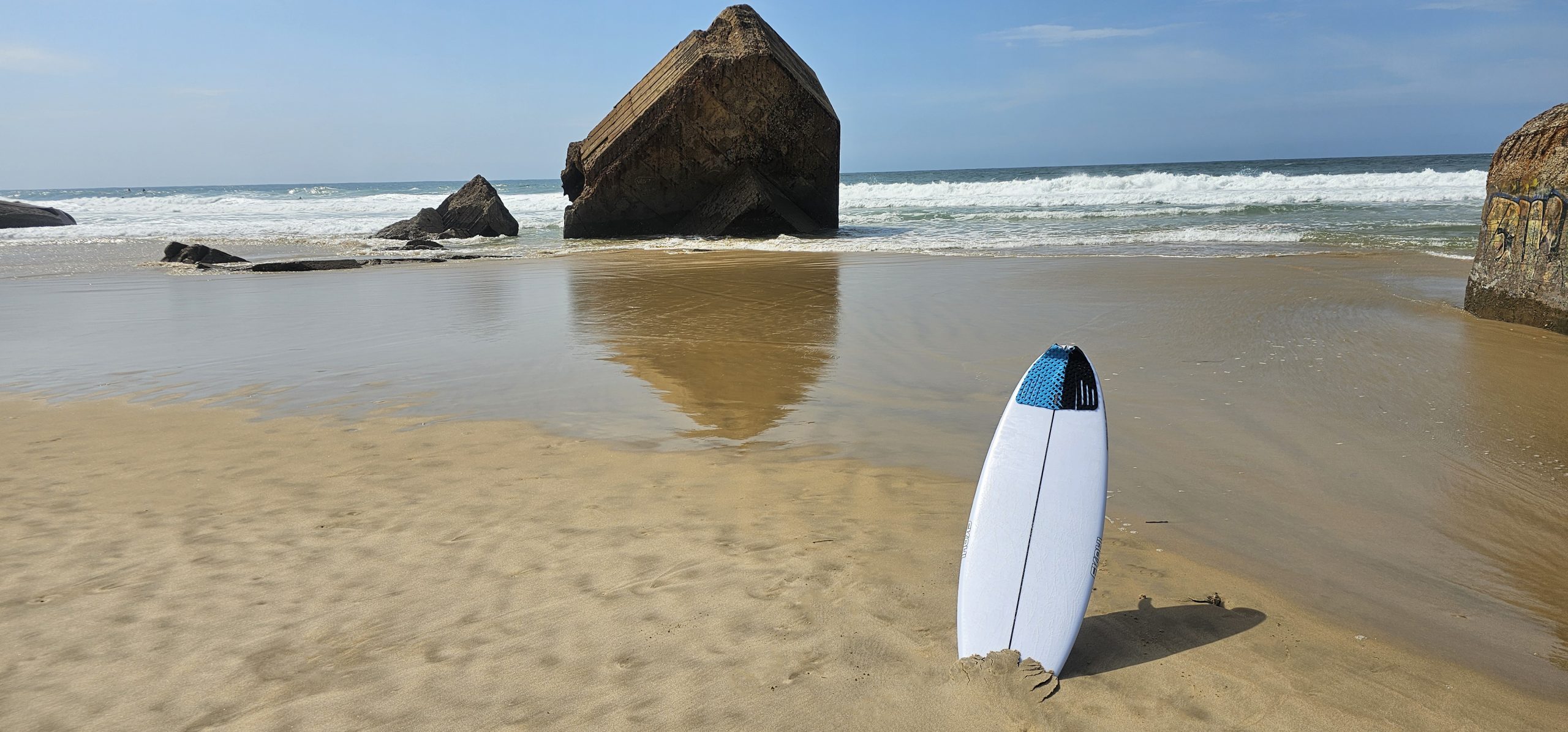 Ekahi surfboard planted upright in the sand on a beach, lifestyle photo with rock formations in the background